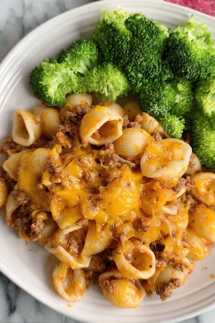 The dish shows a plate filled with three layers: the bottom layer is small pasta shells mixed with cooked ground meat that is soft brown in color; the middle layer is melted cheddar cheese with an orange and golden-brown texture spread evenly over the pasta, giving it a slightly crispy look in places; the top layer is bright green broccoli florets placed on one side of the plate. The plate is white, and the surface beneath is white marble. Photo taken with an iphone --ar 2:3 --v 7 - Cheesy Hamburger Pasta Bake, cheesy hamburger casserole, quick comfort food recipes, easy pasta bake, family-friendly dinner ideas