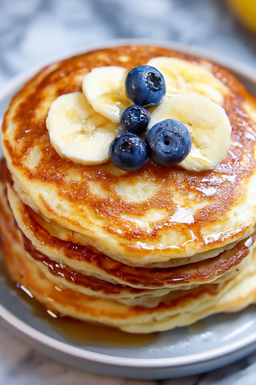 Greek Yogurt Pancakes Recipe 6 A stack of four thick, golden-brown pancakes with a slightly crispy texture on the edges is shown from a close view. On the top pancake, there are three round blueberries placed near the center, glistening slightly, and three slices of pale yellow banana arranged in a row towards the edge. A light drizzle of syrup adds a shiny, wet look on the top pancake. The stack sits on a white plate, all set on a white marbled surface. photo taken with an iphone --ar 2:3 --v 7 - Greek Yogurt Pancakes, fluffy Greek yogurt pancakes, easy breakfast pancake recipes, lemon yogurt pancakes, moist healthy pancakes