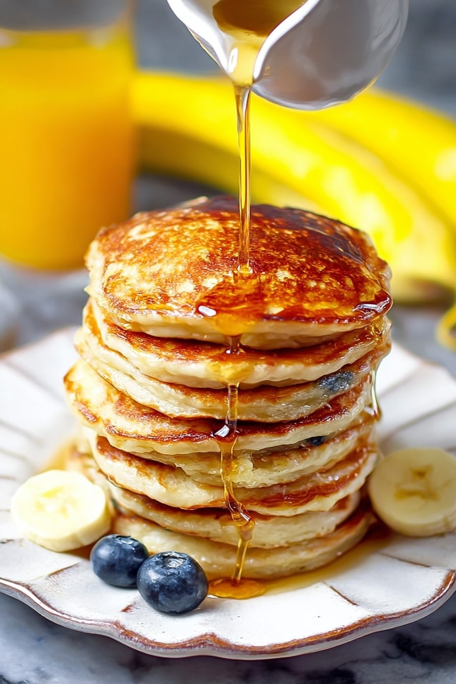 Greek Yogurt Pancakes Recipe 8 A tall stack of seven thick pancakes with a golden brown color is placed on a white plate with a wavy edge. A golden syrup is being poured from a white small pitcher onto the top pancake, flowing down the sides of the stack. Two fresh blueberries sit beside the pancakes on the plate. In the background, blurred bananas and a glass of orange juice are visible on a white marbled surface. The syrup shine and the pancakes’ cooked texture are clearly visible. Photo taken with an iphone --ar 2:3 --v 7 - Greek Yogurt Pancakes, fluffy Greek yogurt pancakes, easy breakfast pancake recipes, lemon yogurt pancakes, moist healthy pancakes