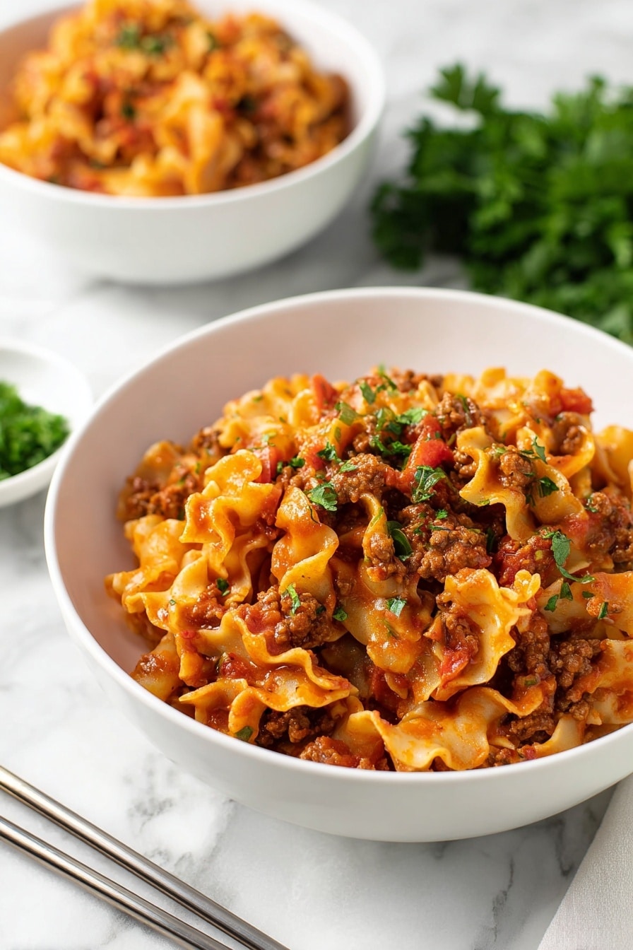 Instant Pot Lazy Lasagna Recipe 8 The image shows a white bowl filled with a layered pasta dish, featuring curly, ruffled-edged noodles mixed with a chunky red tomato sauce and browned ground meat. The pasta is coated in the sauce, giving it a rich orange-red color, with bits of tomato and meat spread evenly throughout. Small green parsley pieces are sprinkled on top for color contrast. Behind the main bowl, there is a second bowl with a similar pasta dish slightly out of focus. The setting includes metal chopsticks resting on a white marbled surface, with some fresh green parsley in the foreground and a blurred bowl of chopped herbs in the background. photo taken with an iphone --ar 2:3 --v 7 - Instant Pot Lazy Lasagna, easy lasagna in Instant Pot, quick lasagna recipe, one-pot lasagna dish, comforting pasta dinner