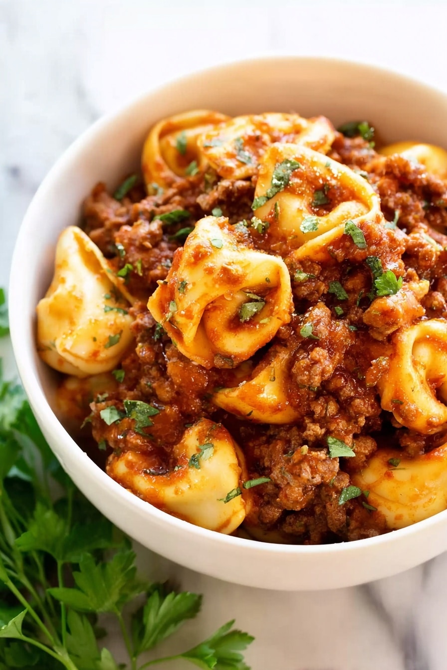 Cheesy Baked Tortellini with Ground Beef Recipe 8 A close-up image of a white bowl filled with three layers of tortellini pasta coated in a rich red meat sauce. The pasta pieces are plump and golden, mixed with browned ground meat and garnished with small green herb leaves, likely parsley. The pasta shapes and meat pieces create a textured look with a mix of rough and smooth surfaces. The bowl is placed on a white marbled surface with a sprig of fresh parsley beside it. Photo taken with an iphone --ar 2:3 --v 7 - Cheesy Baked Tortellini with Ground Beef, baked tortellini recipes, hearty pasta casserole, cheesy ground beef pasta, family-friendly baked pasta