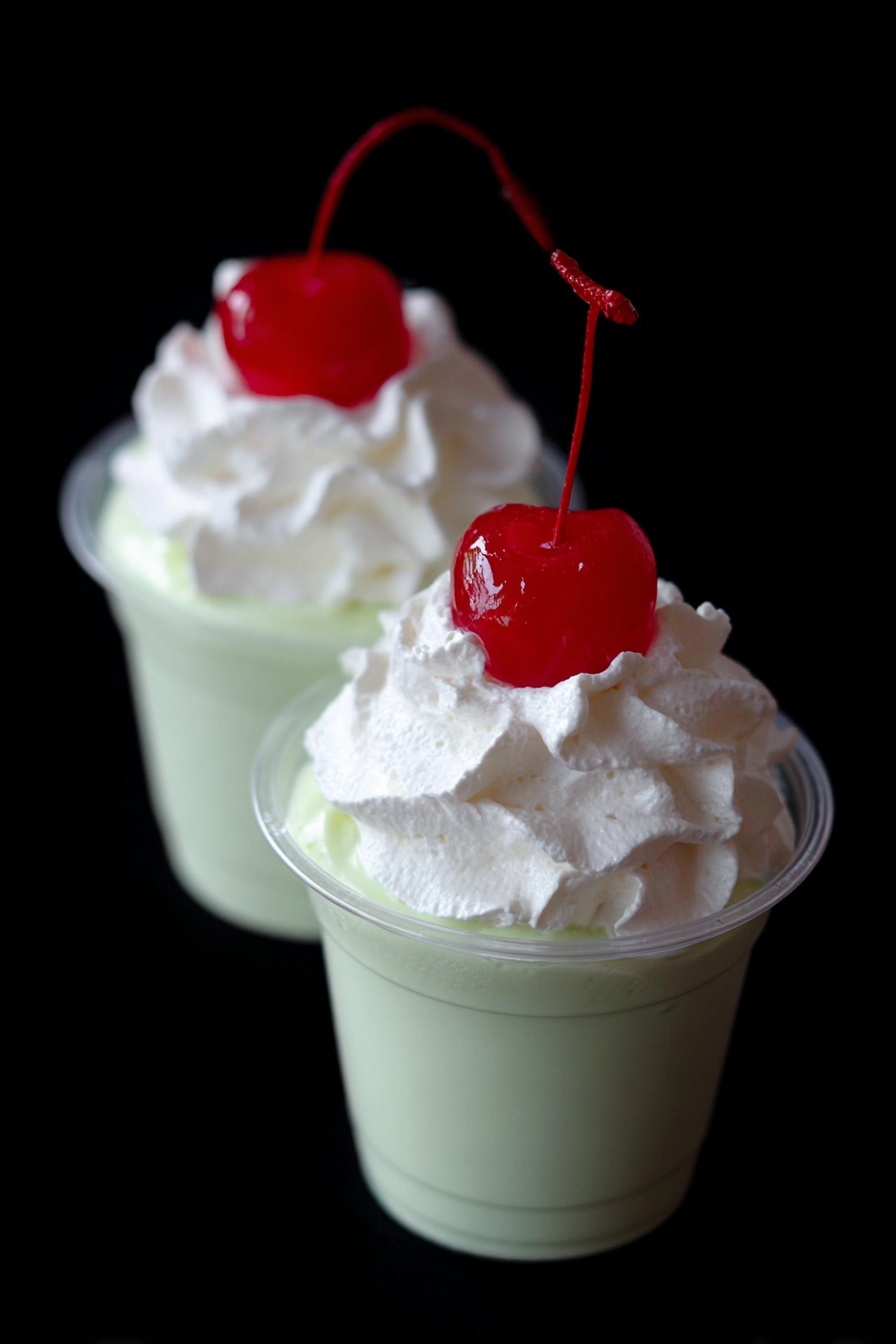Shamrock Shake Pudding Shots Recipe 8 Two small clear cups filled with a smooth light green creamy layer, topped with a thick dollop of white whipped cream. Each cup has a bright red cherry with a long stem placed on top of the whipped cream. The background is black, making the colors of the dessert stand out clearly. The photo taken with an iphone --ar 2:3 --v 7 - Shamrock Shake Pudding Shots, minty green dessert shots, St. Patrick's Day party desserts, boozy pudding shots, festive mint dessert recipes