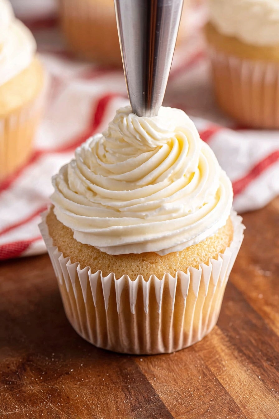 Almond Raspberry Cupcake Delights Recipe 8 The image shows a close-up of a vanilla cupcake placed on a wooden surface with a white paper liner. The cupcake has one thick layer of soft, light yellow cake topped with a large swirl of creamy white frosting that has a smooth, ridged texture and a small peak on top. In the background, there are a couple of similar cupcakes and a white ramekin filled with dark red jam, all placed on the wooden surface with a white marbled texture. Photo taken with an iphone --ar 2:3 --v 7 - Almond Raspberry Cupcake Delights, almond raspberry cupcakes, raspberry-filled cupcakes, almond frosting recipes, berry-filled cupcake ideas