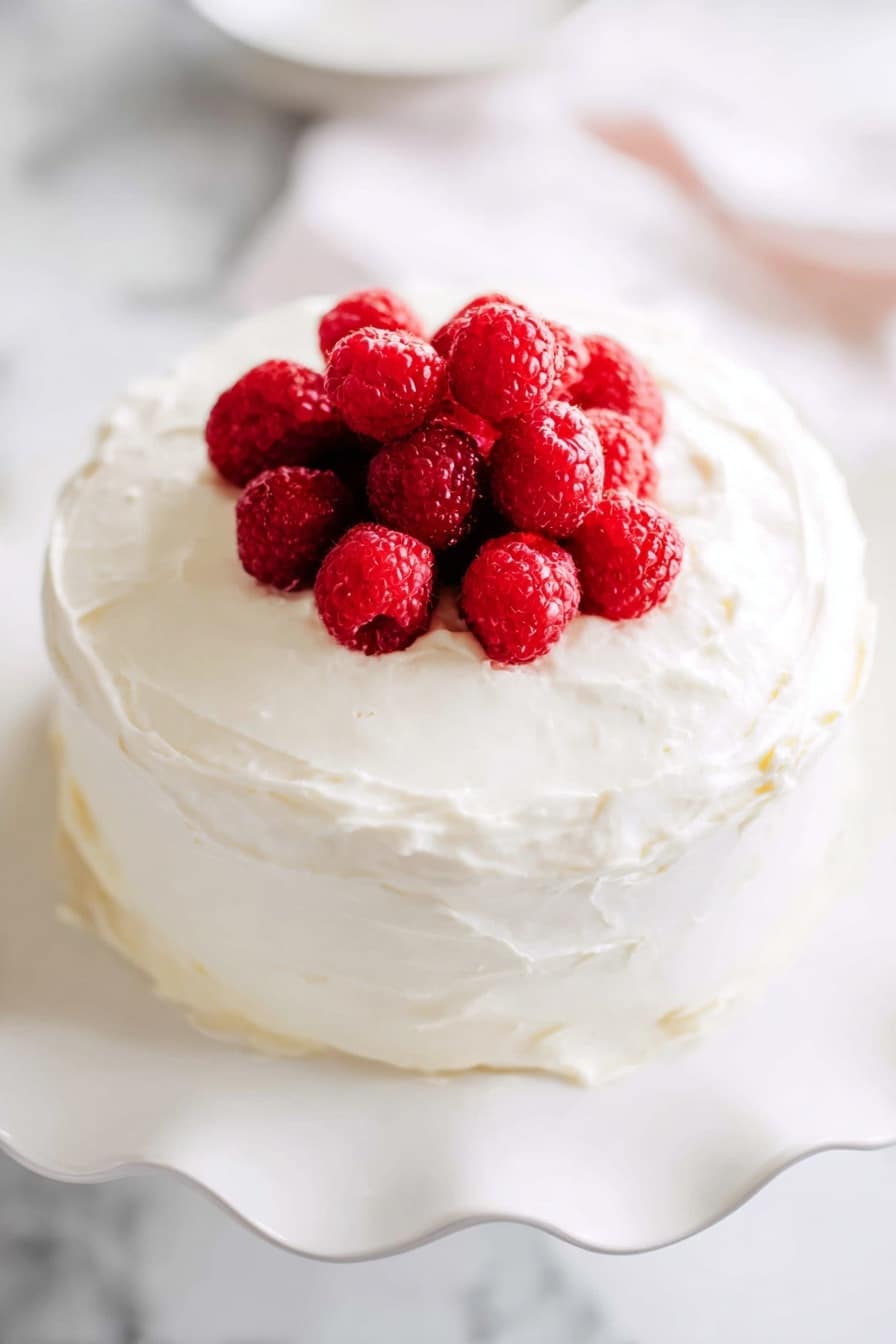 Lemon Raspberry Cake with Cream Cheese Frosting Recipe 6 A round cake with two thick layers covered fully in smooth, white cream frosting is placed on a white plate with wavy edges. On top of the cake is a small pile of fresh, bright red raspberries clustered in the center, adding a pop of color against the white frosting. The cake sits on a white marbled surface with soft, natural light brightening the scene. photo taken with an iphone --ar 2:3 --v 7 - Lemon Raspberry Cake with Cream Cheese Frosting, Lemon Raspberry Cake, Raspberry Layer Cake, Lemon Dessert Recipe, Cream Cheese Frosting Cake