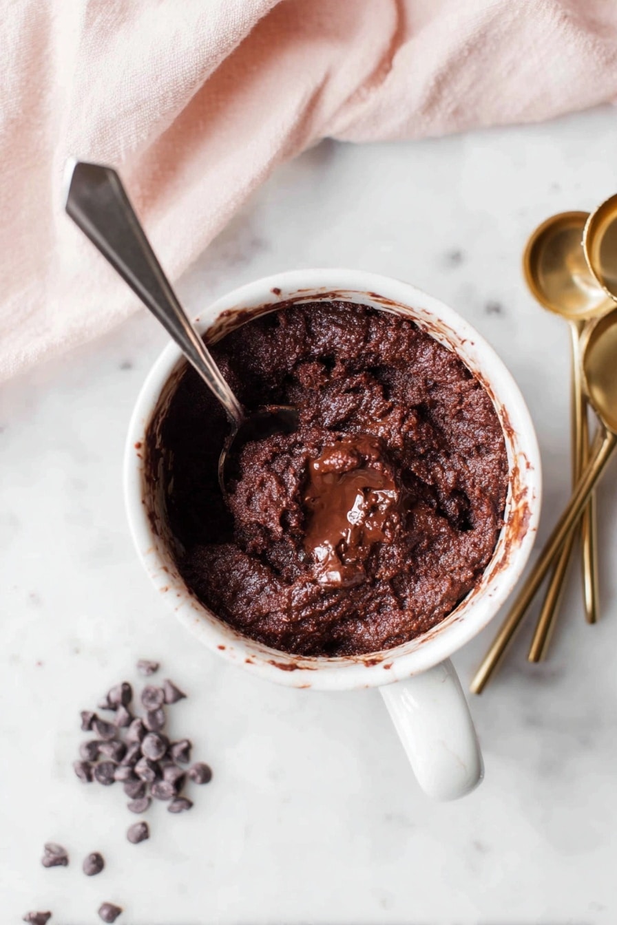 A white mug filled with thick, dark brown chocolate cake batter, showing a rough texture with melted chocolate in the middle, has a silver spoon inside. The mug sits on a white marbled surface with four gold measuring spoons and a small pile of dark chocolate chips nearby. A soft pink cloth is partially visible in the upper left corner. photo taken with an iphone --ar 2:3 --v 7 - Quick Chocolate Mug Cake, easy microwave chocolate cake, solo dessert recipes, fast chocolate mug dessert, simple mug cake treat