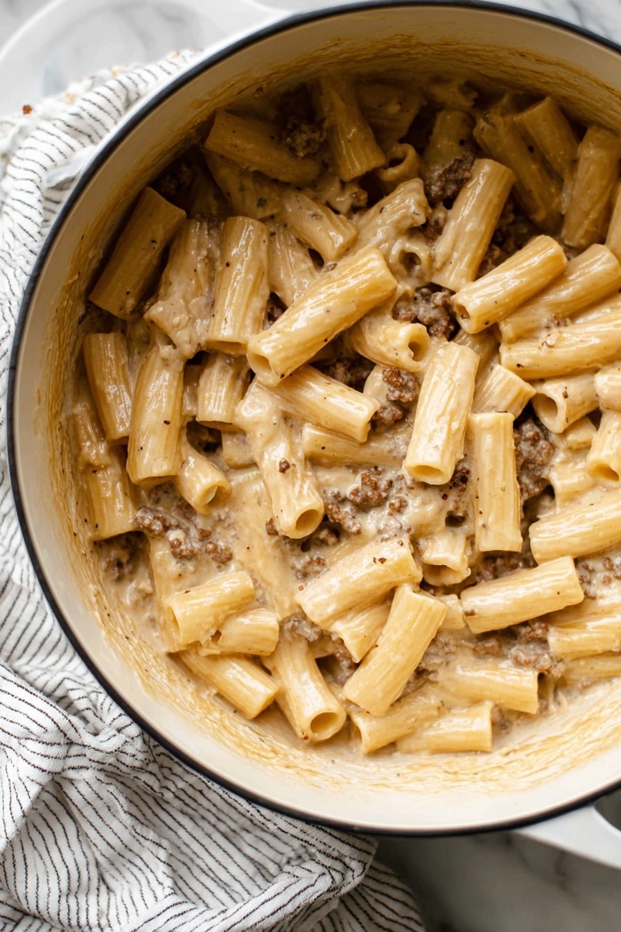 Creamy Beef Pasta Recipe 6 This image shows a close-up of a large white pot filled with creamy pasta. The pasta is tubular and thick, coated in a smooth, light beige sauce with small bits of browned ground meat mixed evenly throughout. The sauce gives the pasta a shiny, rich texture. The pot is set on a white marbled surface with a white and black striped cloth nearby. The edges of the pot have some sauce residue, showing it is freshly stirred. photo taken with an iphone --ar 2:3 --v 7 - Creamy Beef Pasta, creamy beef pasta recipe, hearty beef pasta dish, easy beef pasta dinner, cheesy beef pasta