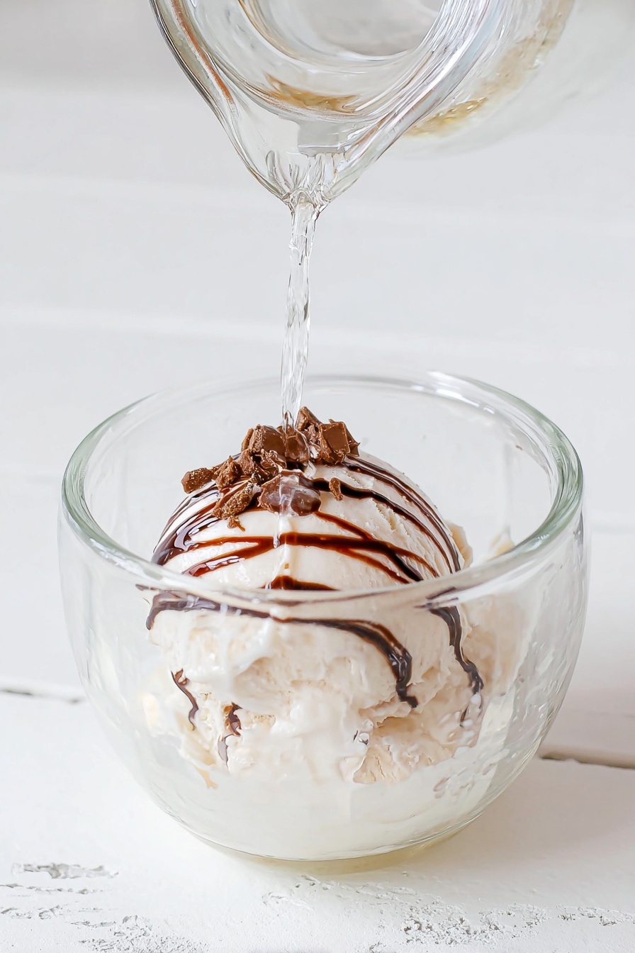 RumChata Hot Cocoa Bombs Recipe 6 A clear glass cup sits on a white marbled surface. Inside the cup, there is a round, white scoop of ice cream with thin chocolate syrup lines drizzled on top. Some small brown chocolate pieces rest on the syrup near the top of the ice cream. Clear liquid is being poured over the ice cream from a glass container above. The light reflects softly off the smooth surface of the ice cream and liquid, creating a fresh and inviting look. photo taken with an iphone --ar 2:3 --v 7 - RumChata Hot Cocoa Bombs, boozy hot chocolate bombs, hot cocoa bombs with RumChata, creamy hot chocolate spheres, alcohol-infused hot cocoa treats