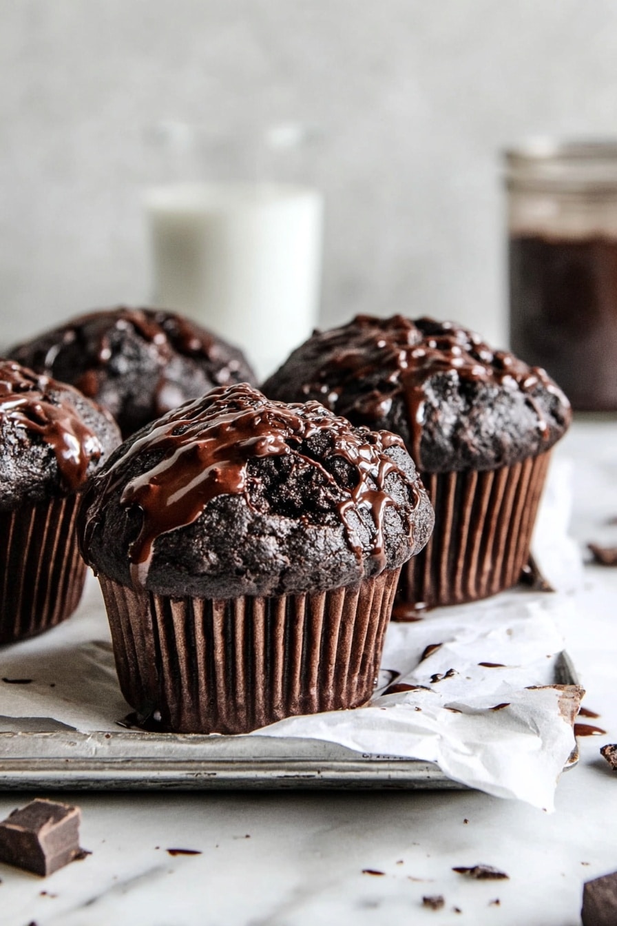 Double Chocolate Chip Muffins Recipe 8 The image shows four rich dark chocolate muffins sitting on a silver tray lined with white parchment paper. Each muffin is topped with glossy dark chocolate drizzle that slightly drips down the sides, adding a shiny texture to the rough cracked tops. The muffins have dark brown, almost black, ridged paper cups. Bits of chocolate pieces and drips of chocolate sauce are scattered around on the white marbled surface beneath the tray. In the background, there is a blurred white tall glass and a jar with dark contents, keeping attention on the muffins in the front. Photo taken with an iphone --ar 2:3 --v 7 - Double Chocolate Chip Muffins, chocolate muffins, moist chocolate muffins, easy chocolate muffin recipe, homemade chocolate muffins