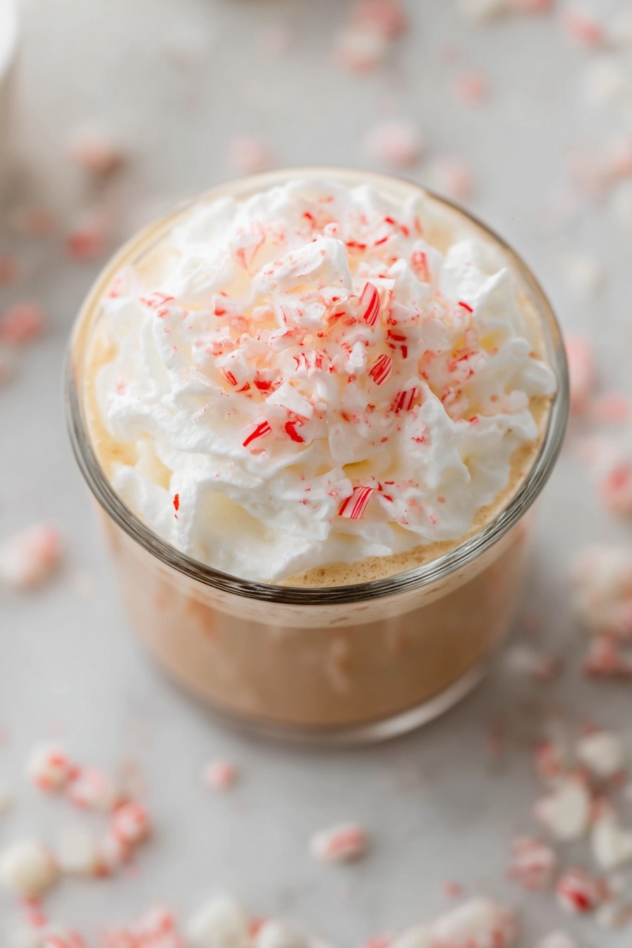 Peppermint White Mocha Recipe 8 A clear glass filled with a drink that has a light brown layer at the bottom, topped with a thick, fluffy white whipped cream layer. On top of the whipped cream, there are small red and white crushed candy pieces spread evenly. The glass sits on a white marbled surface with blurry white and red candy pieces scattered around. The photo is taken from above, showing mostly the whipped cream and candy topping. photo taken with an iphone --ar 2:3 --v 7 - Peppermint White Mocha, peppermint white mocha, white mocha drink, festive coffee recipe, holiday coffee beverages