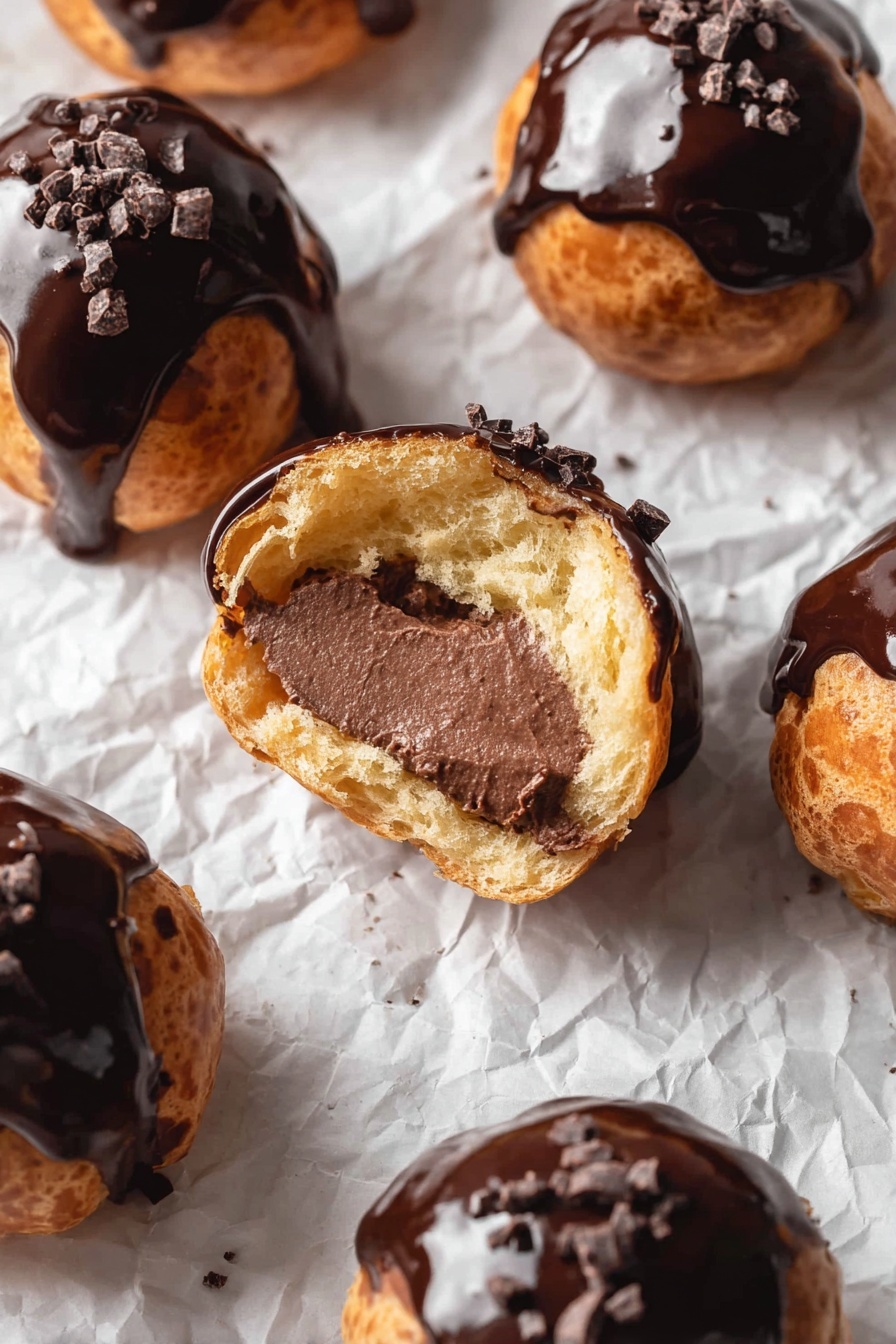 Chocolate Cream Puffs with Pastry Cream Recipe 6 A white plate holds six round pastries, three whole and three cut in half. Each pastry has a golden brown outer layer with a shiny dark chocolate glaze on top, sprinkled with small dark chocolate bits. Inside, the pastries reveal a rich, smooth dark chocolate filling that fills the hollow center. The plate rests on parchment paper over a wooden board, all placed on a white marbled surface. Photo taken with an iphone --ar 2:3 --v 7 - Chocolate Cream Puffs with Pastry Cream, Chocolate Choux Pastry, How to Make Cream Puffs, Homemade Chocolate Filled Pastries, French Cream Puff Dessert