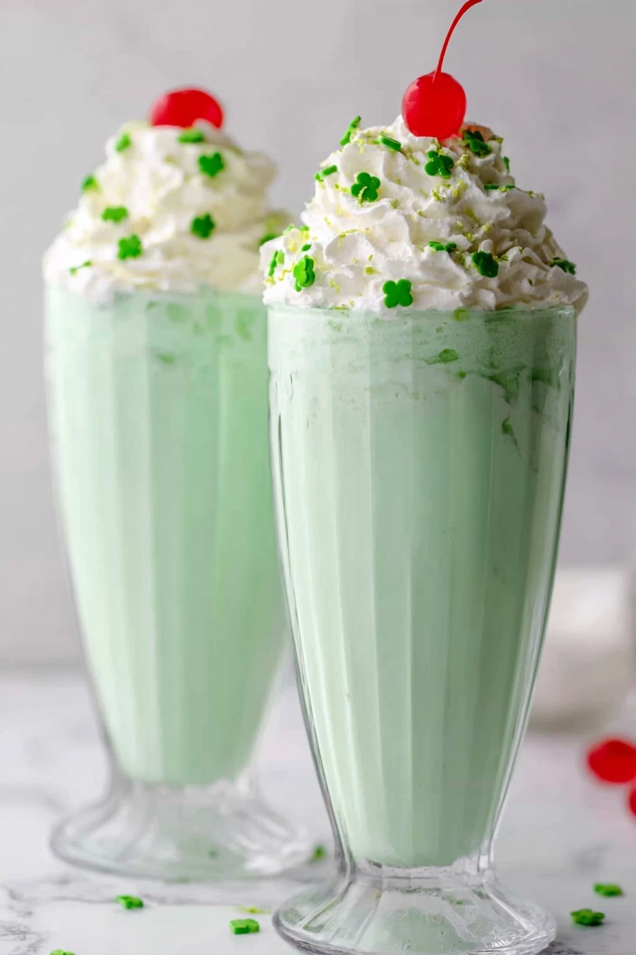 Mint Shamrock Shake Recipe 6 Two tall, clear glasses filled with light green milkshake that looks smooth and creamy stand side by side on a white marbled surface. Each glass has a generous swirl of white whipped cream on top, decorated with small green clover-shaped sprinkles scattered around and a bright red cherry placed at the peak. The light green milkshake is thick and fills most of the glass, which has a ridged pattern that adds texture. The background is simple and softly blurred, keeping the focus on the milkshake glasses. Photo taken with an iphone --ar 2:3 --v 7 - Mint Shamrock Shake, mint shamrock shake recipe, green mint milkshake, St. Patrick's Day shamrock shake, homemade mint shake