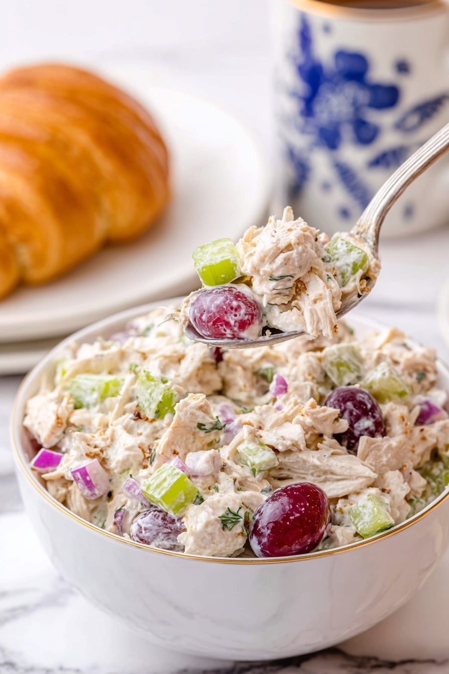 Chicken and Grape Salad Recipe 6 A white bowl filled with a creamy chicken salad showing shredded light-colored chicken mixed with light green celery pieces, small purple-red grape halves, small bright purple onion chunks, and some specks of herbs. A silver spoon is lifting a spoonful of the chicken salad above the bowl, highlighting the colorful mix. In the background, there is a white cup with blue floral designs and a white plate holding a golden croissant, all placed on a white marbled surface. photo taken with an iphone --ar 2:3 --v 7 - Chicken and Grape Salad, Chicken and Grape Salad Ingredients, Easy Chicken and Grape Salad, Healthy Chicken Grape Salad, Quick Chicken Salad with Grapes