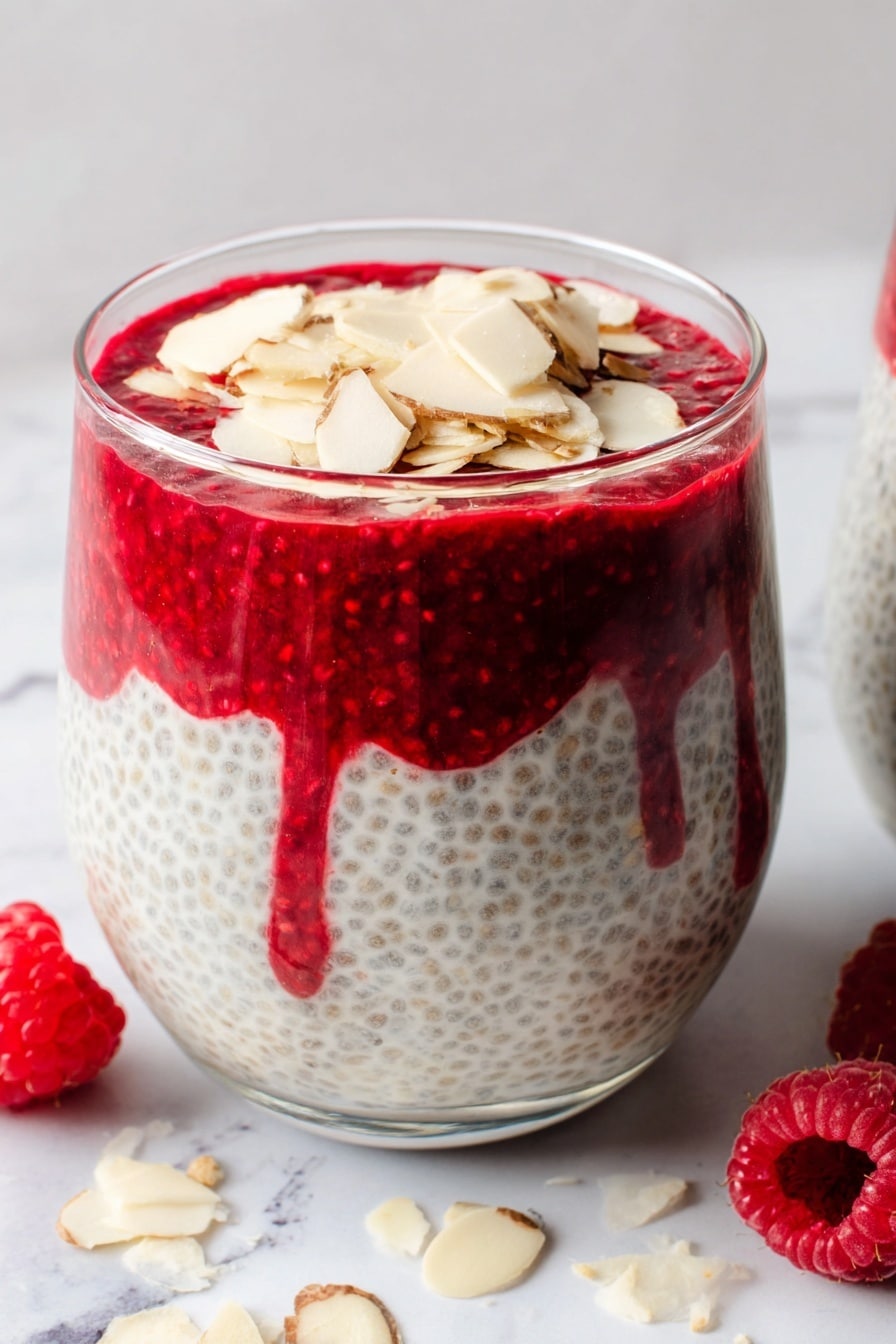 Das Bild zeigt ein klares Glas, das mit drei sichtbaren Schichten gefüllt ist. Die unterste Schicht ist ein cremiger Chia-Pudding mit sichtbaren Chiasamen, hellgrau mit kleinen dunklen Punkten und einer glatten Textur. Darüber befindet sich ein dickes, leuchtend rotes Himbeerpüree, das leicht in die Chiaschicht tropft und eine reiche und leicht klumpige Textur hat. Die oberste Schicht besteht aus dünn geschnittenen Mandelflocken, blass beige, die locker über das Himbeerpüree verstreut sind. Das Glas steht auf einer weiß marmorierten Oberfläche mit einigen Mandelflocken darüber und zwei frischen Himbeeren an der Seite. Foto aufgenommen mit einem iPhone --AR 2:3 --v 7 - Himbeer-Chia-Hüttenkäse-Pudding, gesunder Frühstückspudding, eiweißreiches Obstdessert, einfache Chia-Pudding-Rezepte, fettarme Snackideen