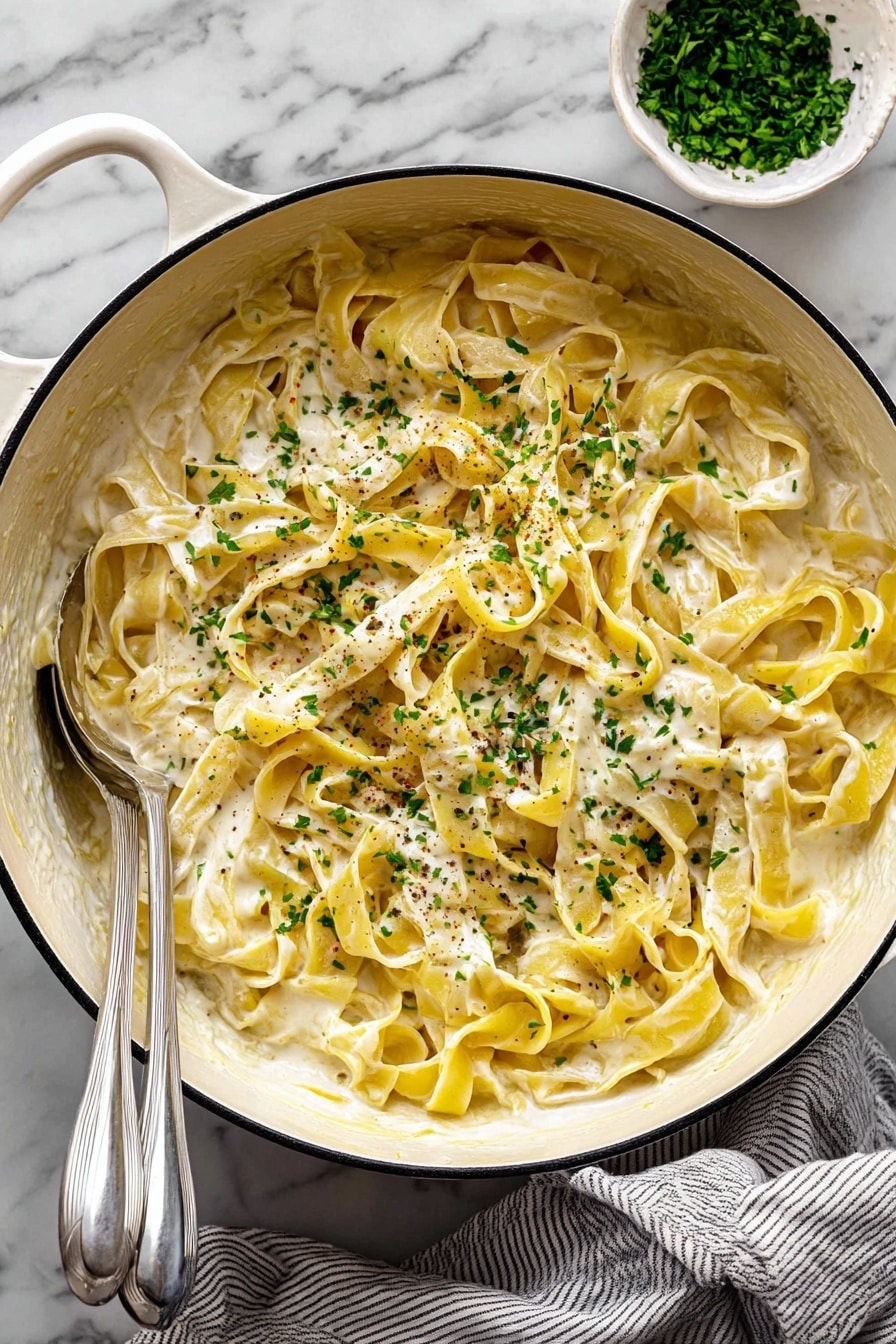 A large white pot filled with creamy yellow pasta ribbons, layered loosely and covered evenly in a smooth, white cream sauce. The pasta is sprinkled with small chopped green herbs and tiny specks of black pepper across the top. Two silver utensils rest inside the pot on the left side, partially covered by the pasta. In the top right corner, there is a small white bowl filled with more green herbs. The pot sits on a white marbled surface with a gray and white striped cloth partially under it. Photo taken with an iphone --ar 2:3 --v 7 - Creamy Lemon Pasta, easy lemon pasta, quick lemon pasta recipe, creamy pasta with lemon, family-friendly lemon pasta